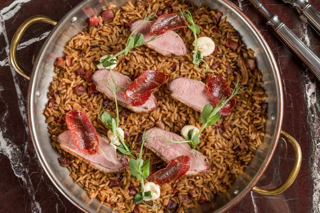 Arroz de jambalaya com fatias de pato, fatias de aipo, microverdes e pedaços de linguiça defumada, servido em uma panela de metal com alças de ouro