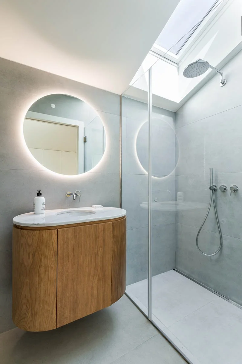 Banheiro moderno com espelho circular com iluminação, pia de madeira com cuba embutida, sabonete líquido e saboneteira, parede de vidro que separa o chuveiro, teto com janela de telhado, chuveiro de teto e controle de água na parede.