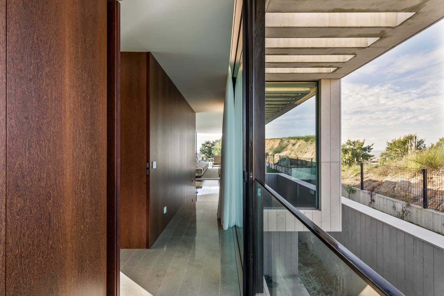 Corredor de um apartamento com parede de madeira, vidro e vista para uma área externa com céu e árvores.