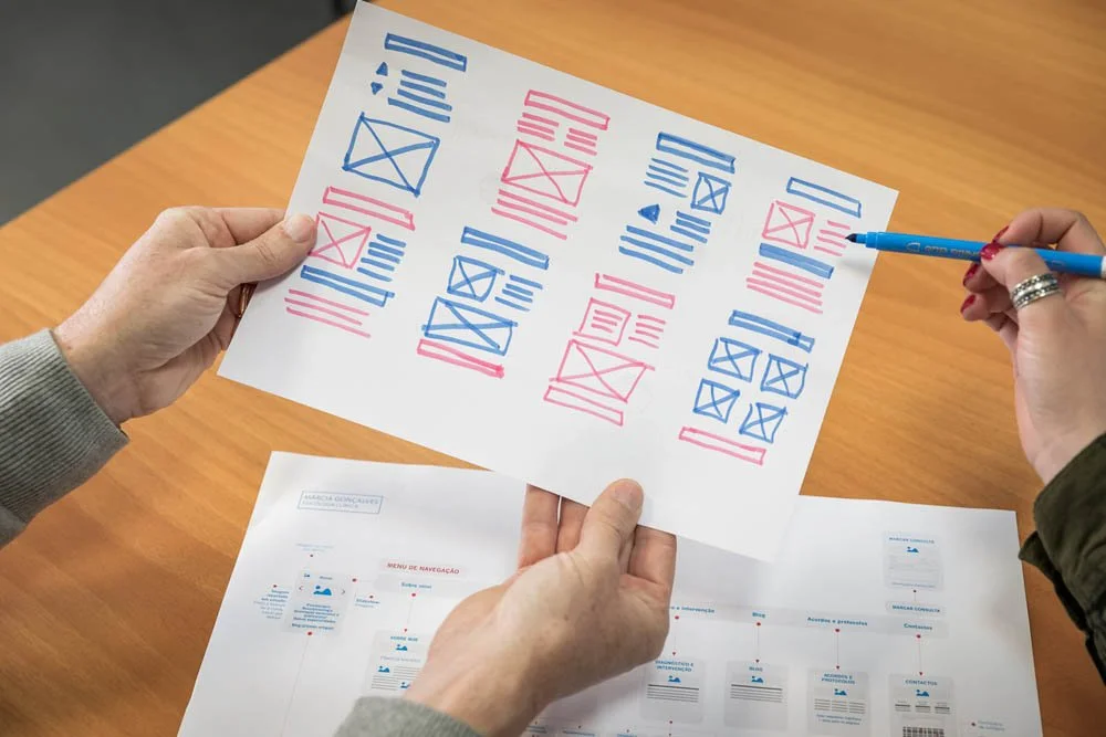 Pessoa segurando uma folha com esboços de layout de site feitos com canetas azul e rosa. Outras folhas com diagramas estão sobre uma mesa de madeira.