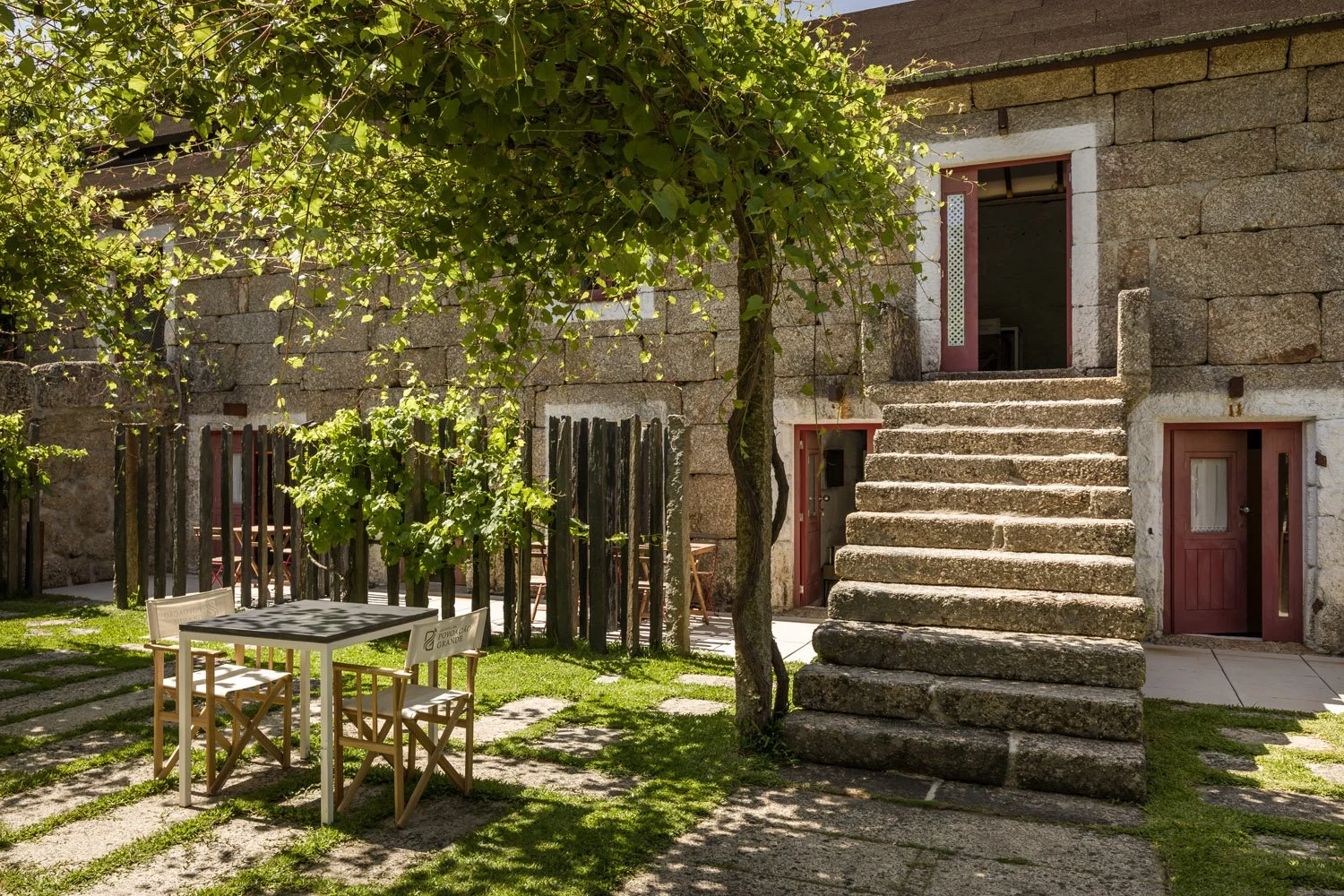 Casa de pedra com escada de pedra, jardim com mesa e cadeiras de madeira, árvores e sombra natural.