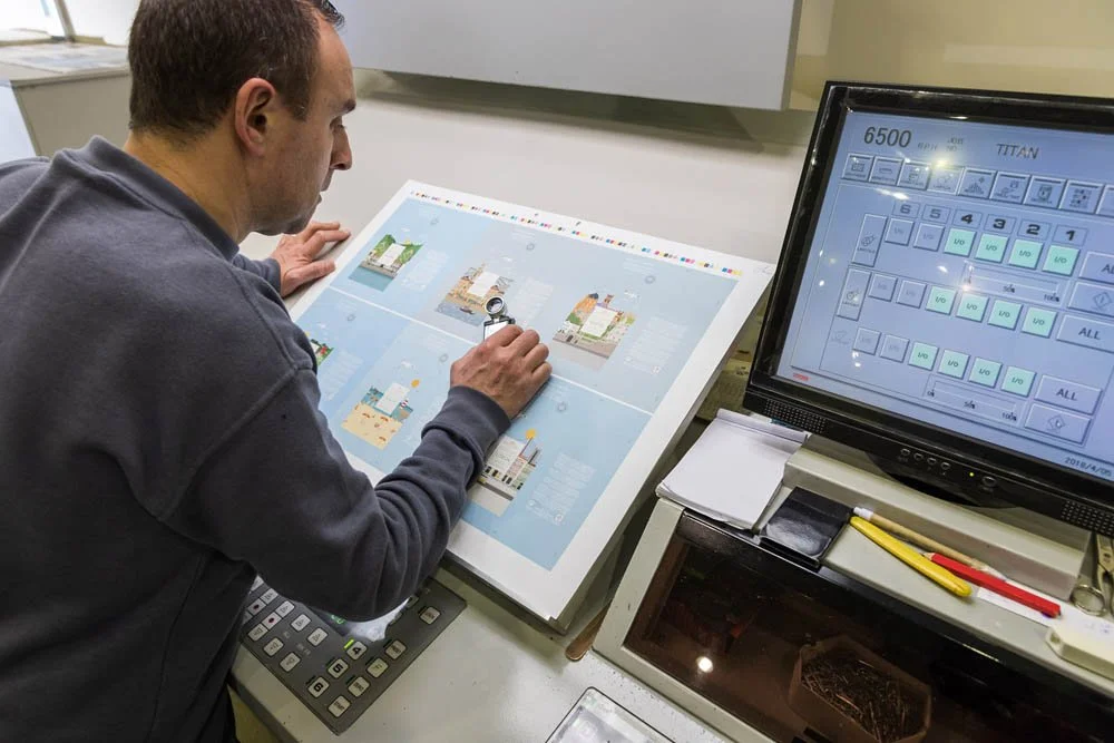 Homem trabalhando com uma mesa digital interativa, usando uma lupa para ampliar a tela, ao lado de um monitor de computador exibindo layout de produção ou controle de estoque, em um escritório.