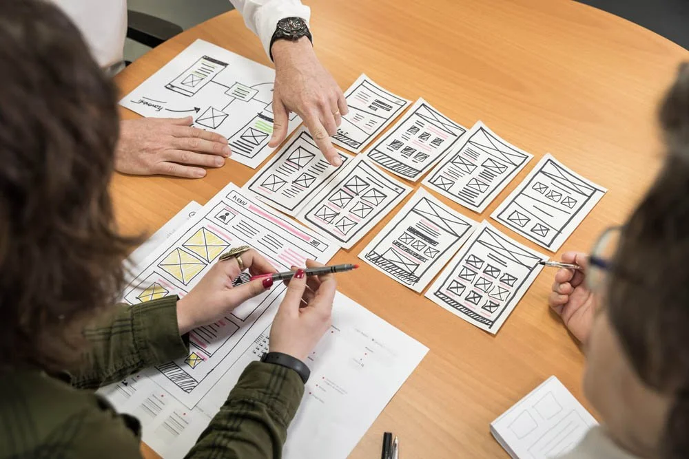 Pessoas discutindo e analisando esboços de design de aplicativos ou páginas web em uma mesa de reunião.