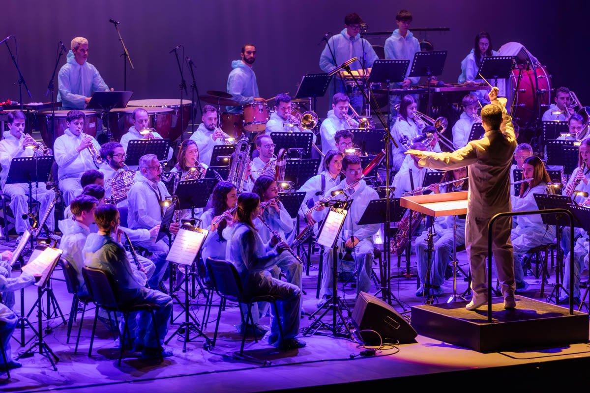 Orquestra de músicos em apresentação, com maestro conduzindo, sob iluminação colorida em palco.