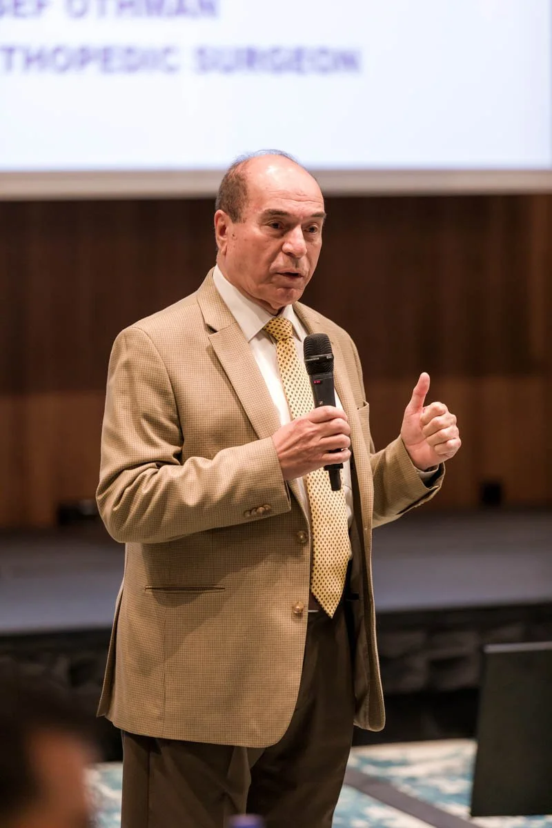 Homem fazendo uma palestra, segurando um microfone, vestindo terno bege e gravata, em um ambiente de conferência.