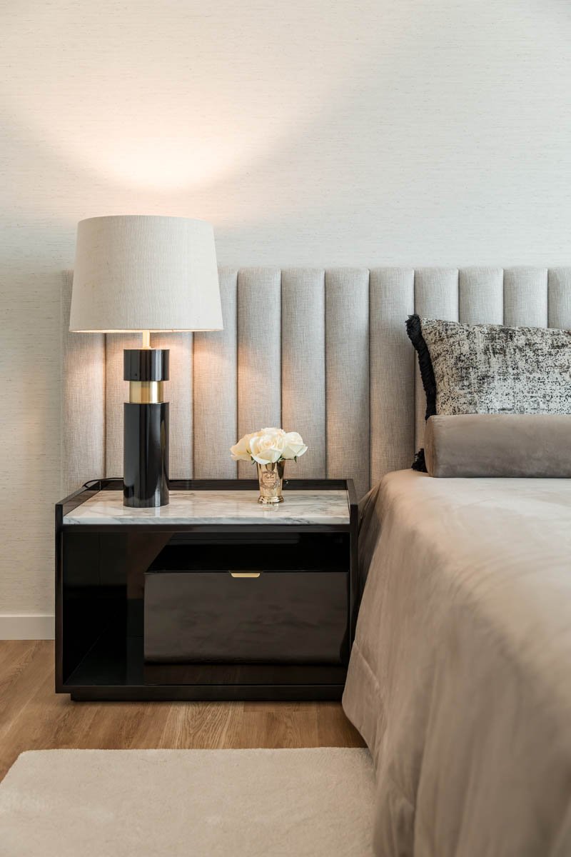 Cama de casal com cabeceira almofadada vertical, travesseiro decorativo cinza e roupa de cama bege, com uma luminária de mesa ao lado, sobre uma cômoda preta com tampo de mármore, decorada com um vaso de flores brancas