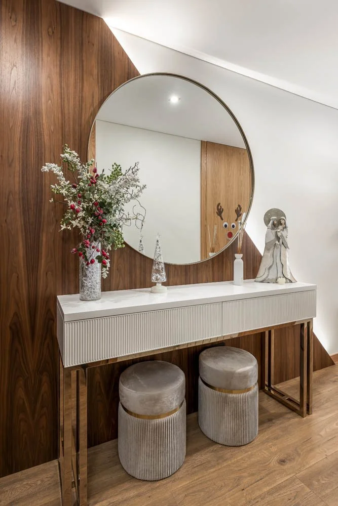 Espelho grande redondo acima de um móvel branco com tampos decorados com vasos, enfeites natalinos e uma escultura de Natal, com dois banquinhos de veludo cinza e detalhes dourados em madeira.