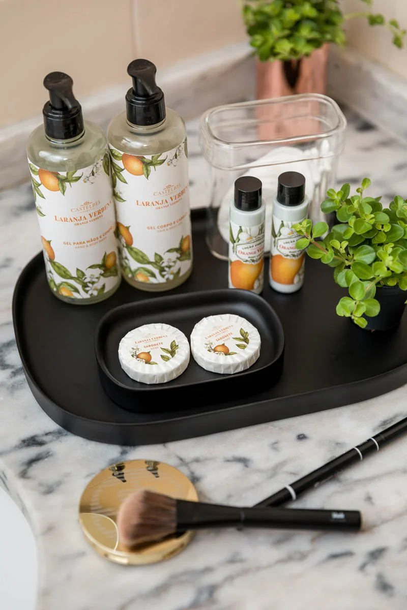 Conjunto de produtos de cuidado pessoal com tema de laranja, incluindo gel para mãos e corpo, sabonetes, produtos em miniatura, uma planta verde, uma escova de maquiagem dourada, uma base e uma caneta. Itens dispostos sobre uma bandeja preta em uma bancada de mármore.