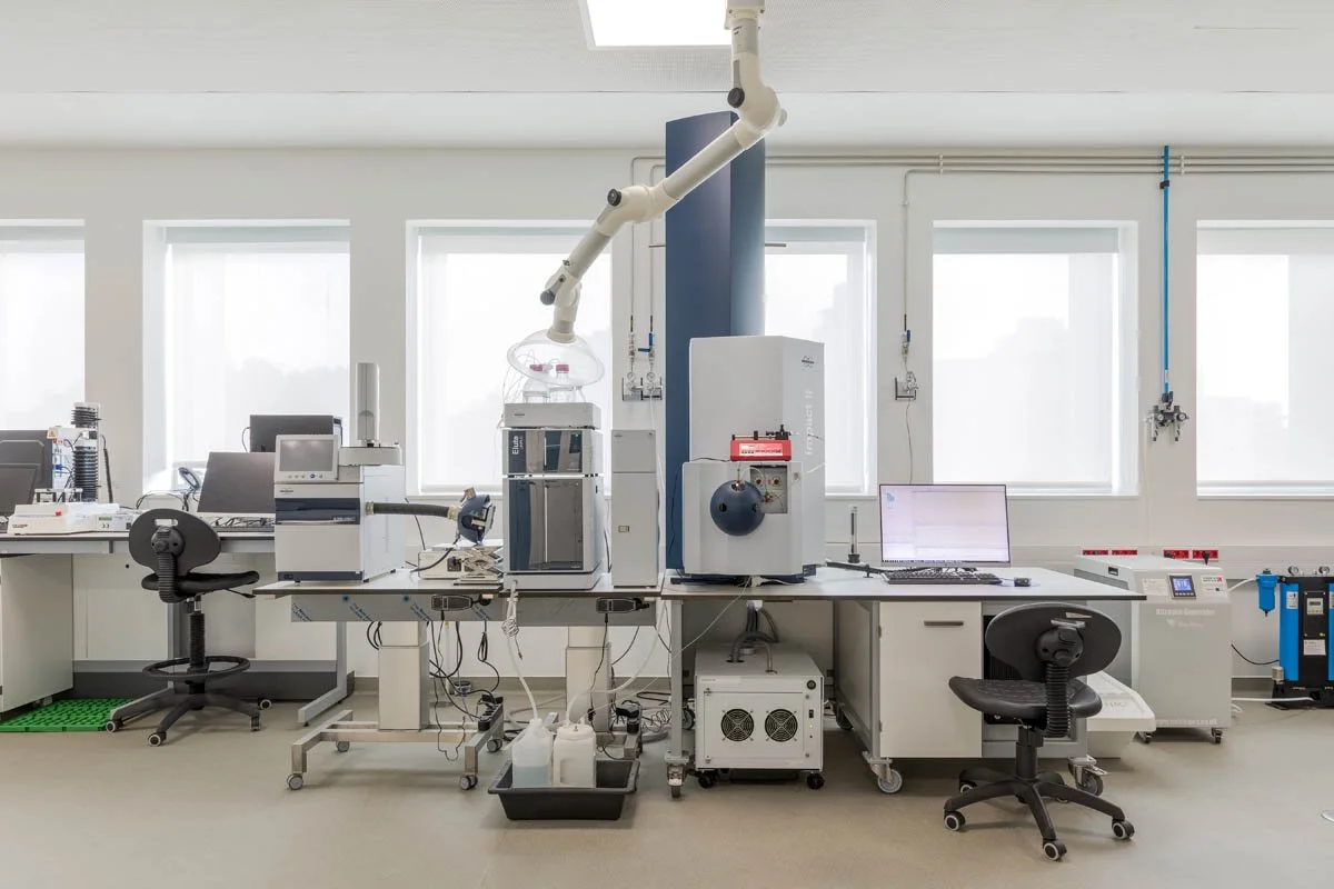 Laboratório com equipamentos de análise científica, incluindo instrumentos de alta tecnologia, mesas de trabalho, computadores e cadeiras, em um ambiente bem iluminado com janelas grandes.