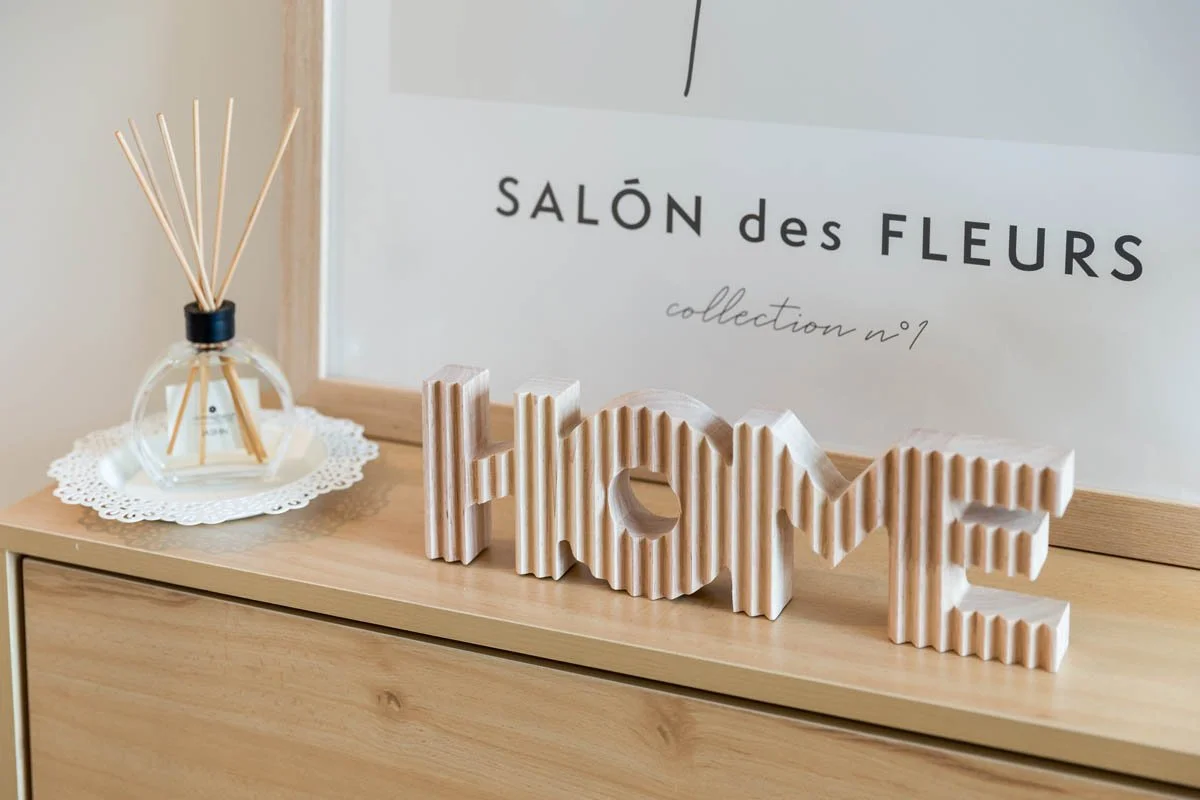 Decoração com uma palavra escrita em madeira que diz 'HOME' e um recipiente de vidro com difusor de aromas ao lado, em cima de uma bancada de madeira, com uma moldura ao fundo com escrito em francês 'salon des fleurs'.