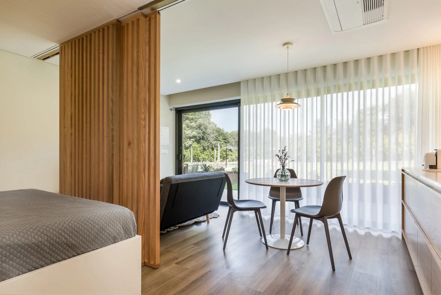 Interior de um apartamento com quarto parcialmente visível, espaço de convivência com uma mesa de jantar, cadeiras, um sofá, cortinas de correr com vista para um jardim externo, piso de madeira.