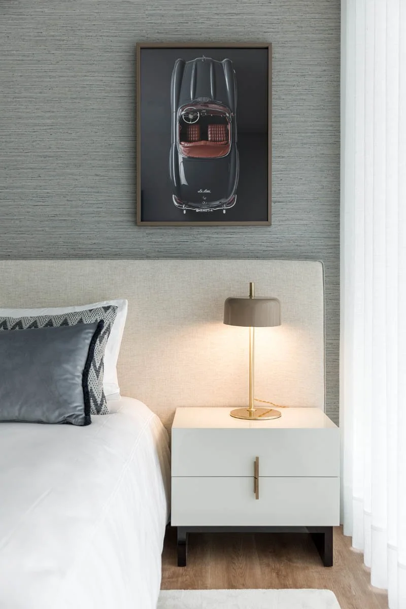 Quarto com cama de cabeceira bege, travesseiro cinza e almofada decorativa, luminária de mesa ao lado da cama, parede de textura cinza, quadro de carro antigo preto na parede, cortina branca na janela ao lado direito.