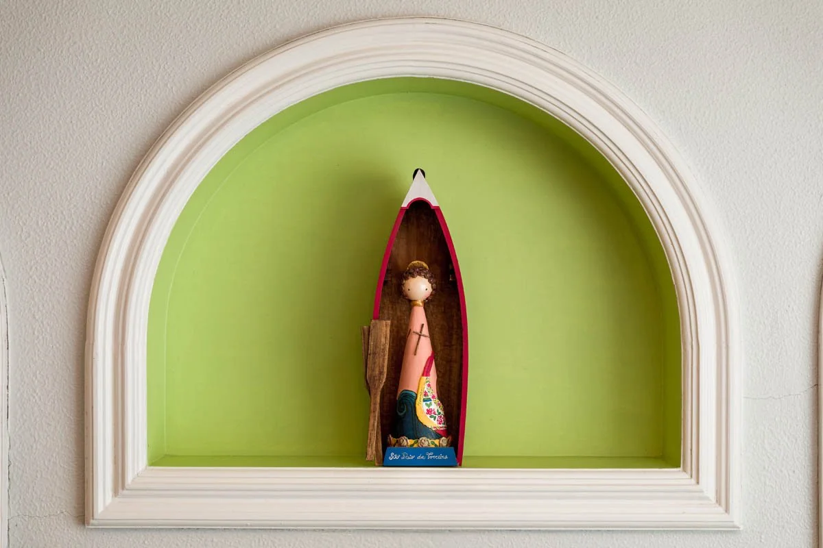 Figura decorativa de uma mulher com roupas coloridas, colocada dentro de uma pequena embarcação, exibida em uma parede com nicho em relevo branco com fundo verde