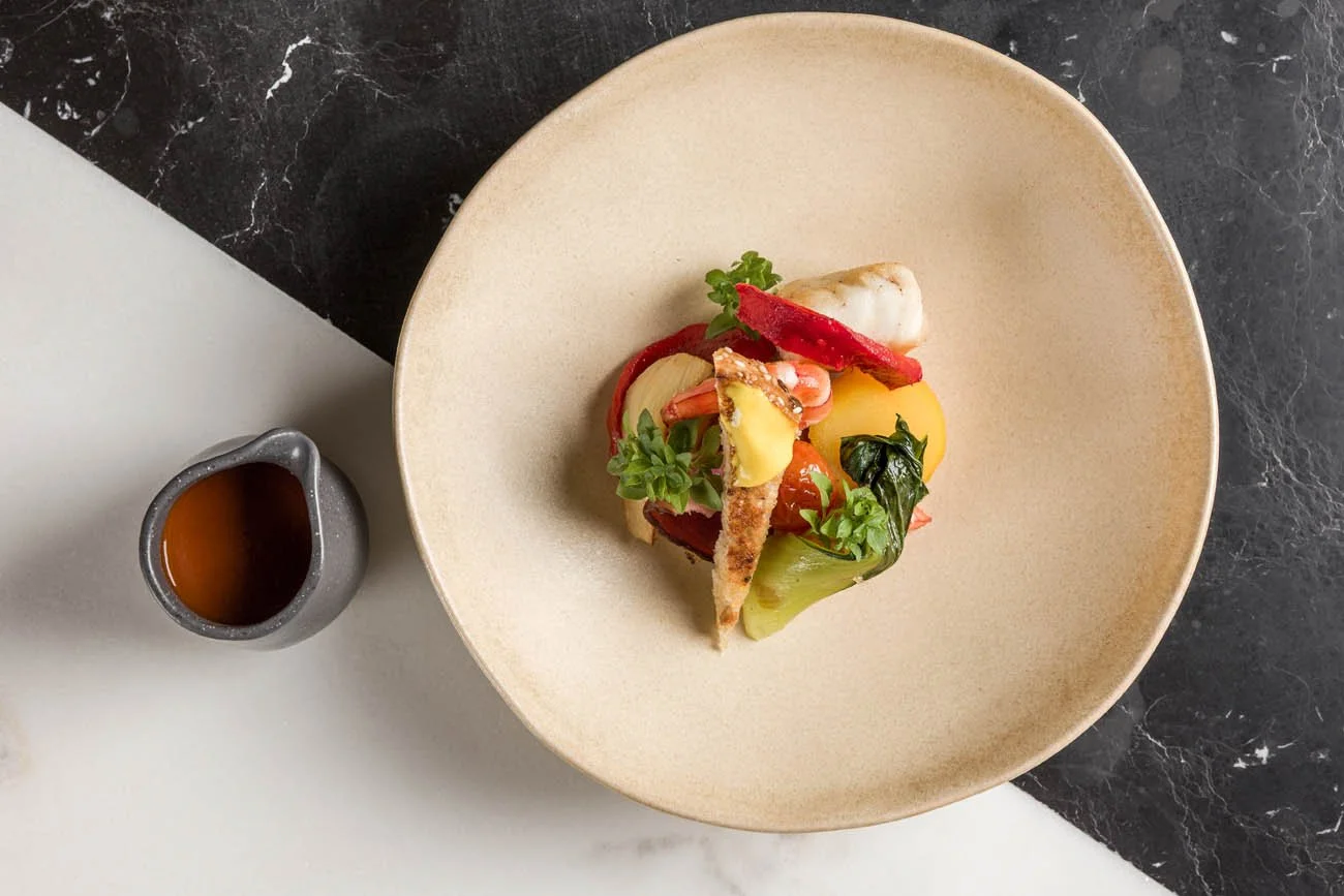 Prato com peixe ao lado de molho escuro, disposto em um prato de cerâmica sobre uma mesa com fundos de mármore branco e preto.