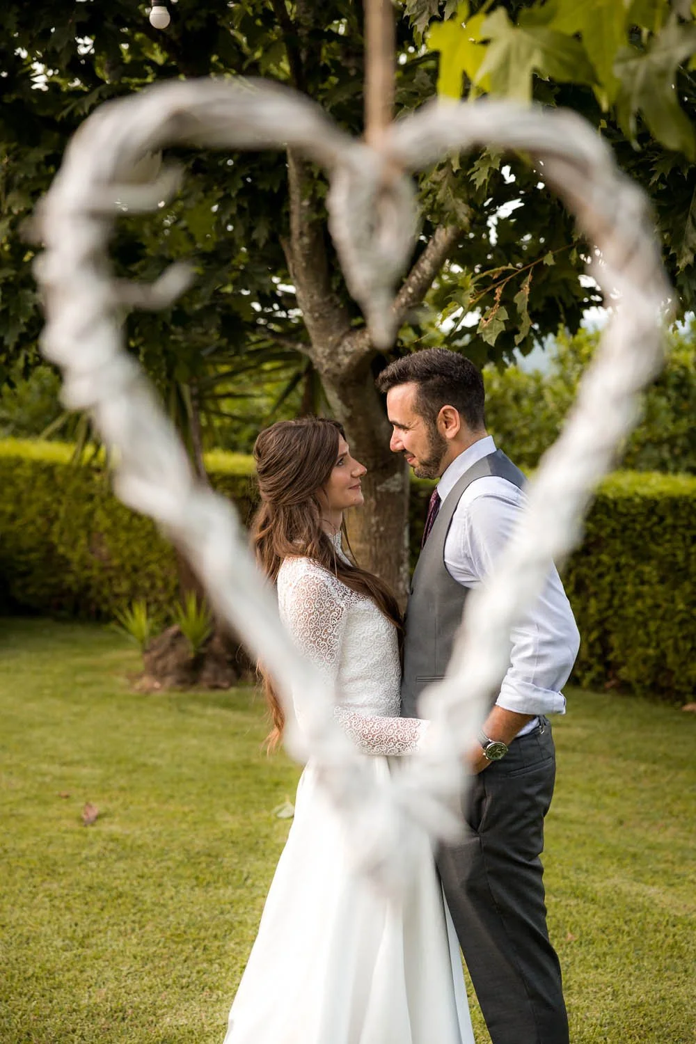 Casal em traje de casamento olhando um para o outro ao ar livre, com árvores ao fundo, vista através de um coração feito com uma estrutura de metal branca.