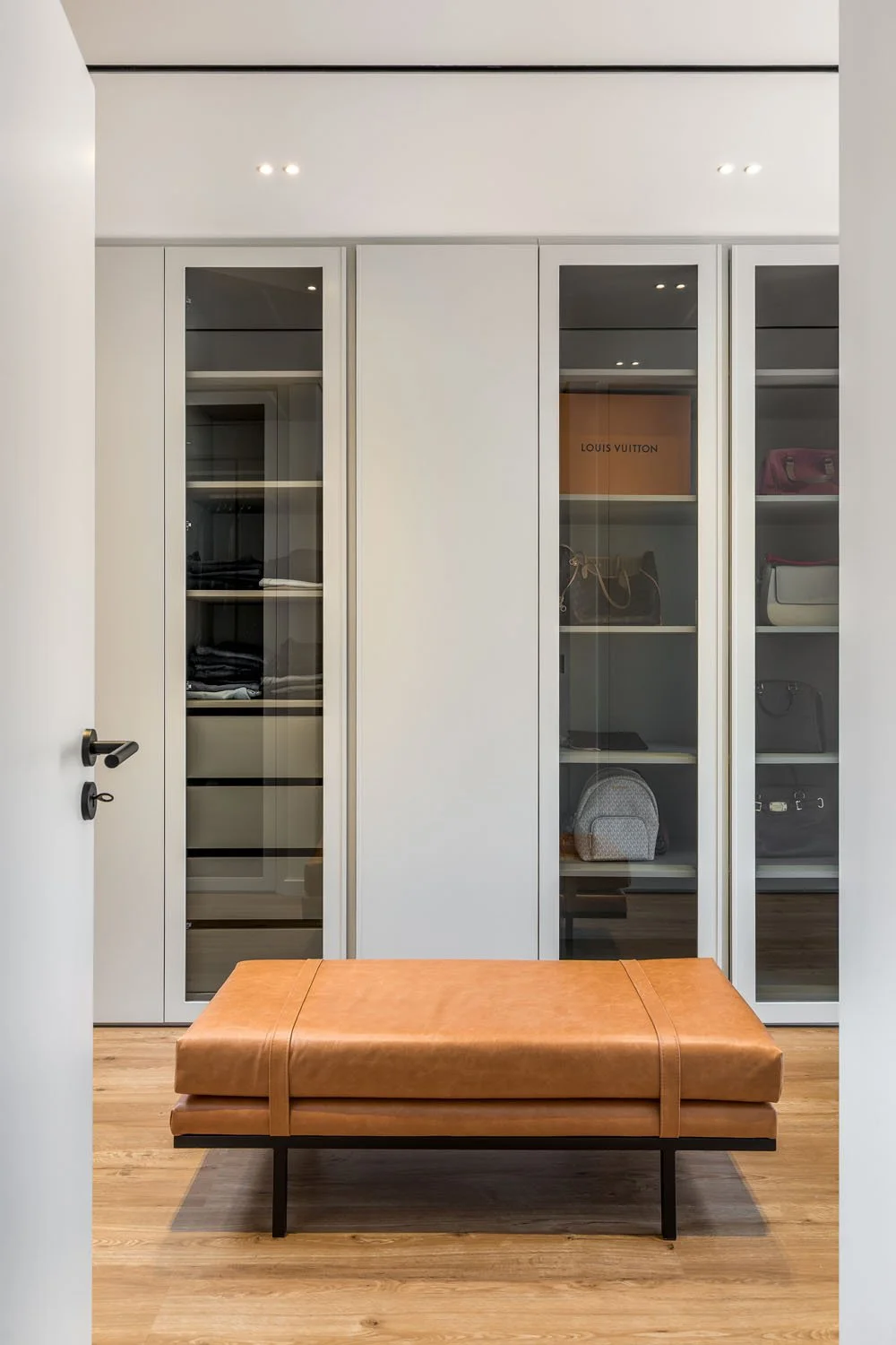 Entrando em um closet com prateleiras de vidro e bolsas de luxo, com um banco de couro marrom na frente e piso de madeira.