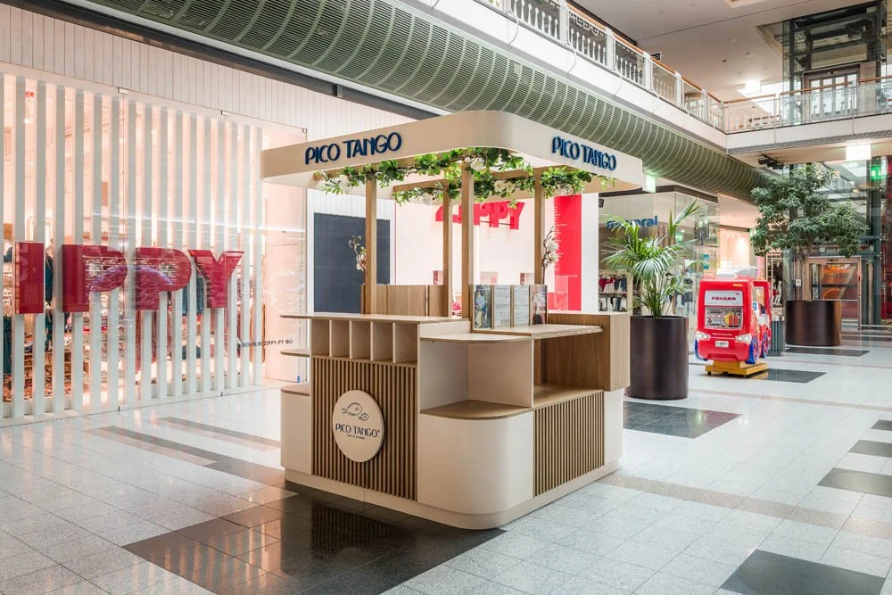 Quiosque com sinalização 'Pico Tango' dentro de um shopping, decorado com plantas e poltronas de madeira, ao lado de uma parede de vidro com letras vermelhas 'P P Y'.