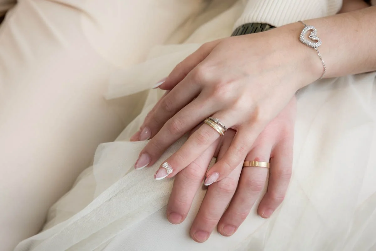 Mãos de um casal com alianças de casamento, mostrando joias e unhas decoradas.