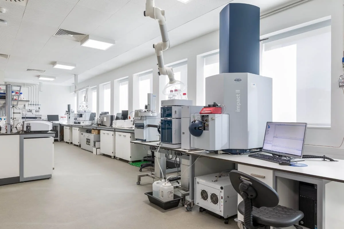 Laboratório de alta tecnologia com equipamentos de análise científica, incluindo computadores, instrumentos laboratoriais e bancos de trabalho, iluminado por luz natural das janelas.