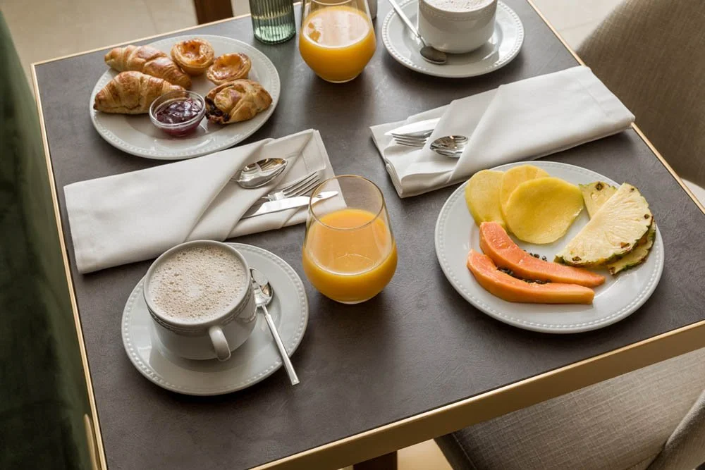 Mesa de café da manhã com croissants, geleia, suco de laranja, café com espuma, ovos, queijo, salame e frutas.