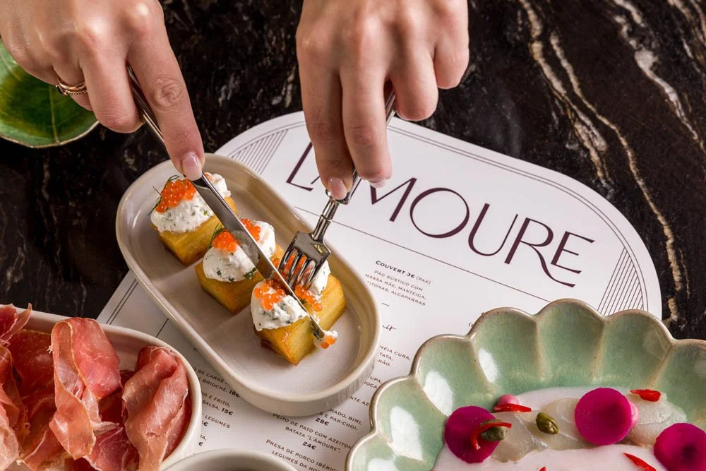 Pessoa cortando petiscos em um prato de porcelana branca, com diversos pratos de comida ao redor, incluindo presunto e uma salada decorativa