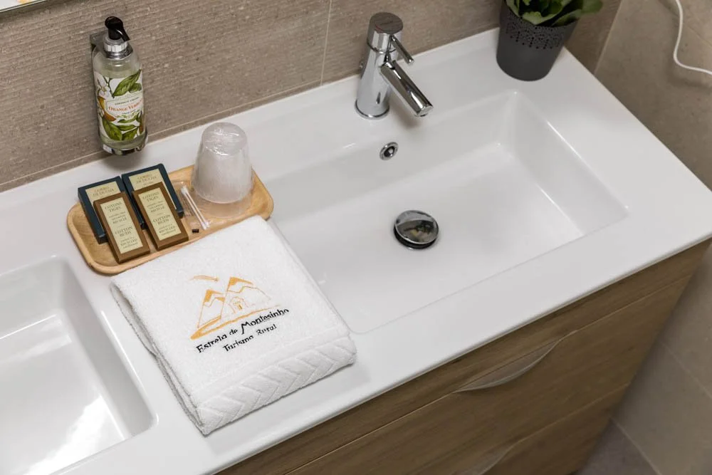 Pia de banheiro com sabonete líquido, copo de vidro, toalha branca com o texto e símbolo de hospedagem, e uma garrafinha de sabonete de aroma de ervas ao lado da pia, além de uma planta no canto superior direito.
