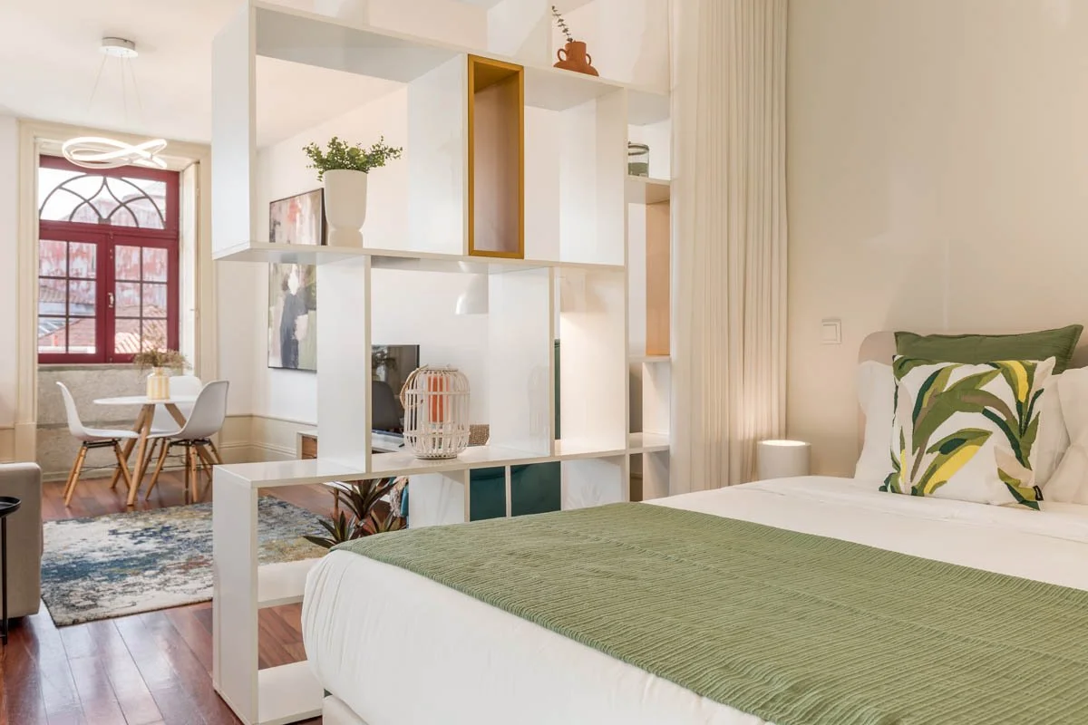 Quarto com cama de casal com roupa de cama branca e travesseiros com estampa de folhas verdes, divisória de estantes, janela com cortinas brancas e área de jantar com mesa e cadeiras brancas ao fundo.