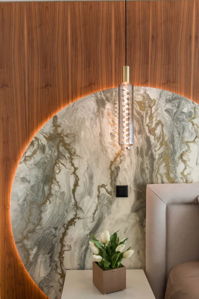 Iluminação decorativa com lâmpada pendente sobre parede de mármore e madeira, com vaso de tulipas brancas na mesa ao lado da cama.