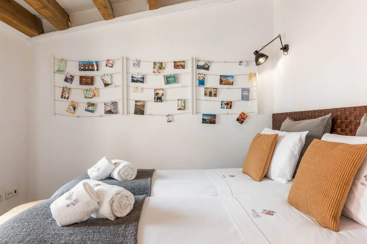 Cama de casal com travesseiros cinza e amarelos, toalhas brancas e uma colcha cinza, em um quarto com parede branca, parede de madeira e teto de madeira exposta, decorado com fotos penduradas por fios na parede.