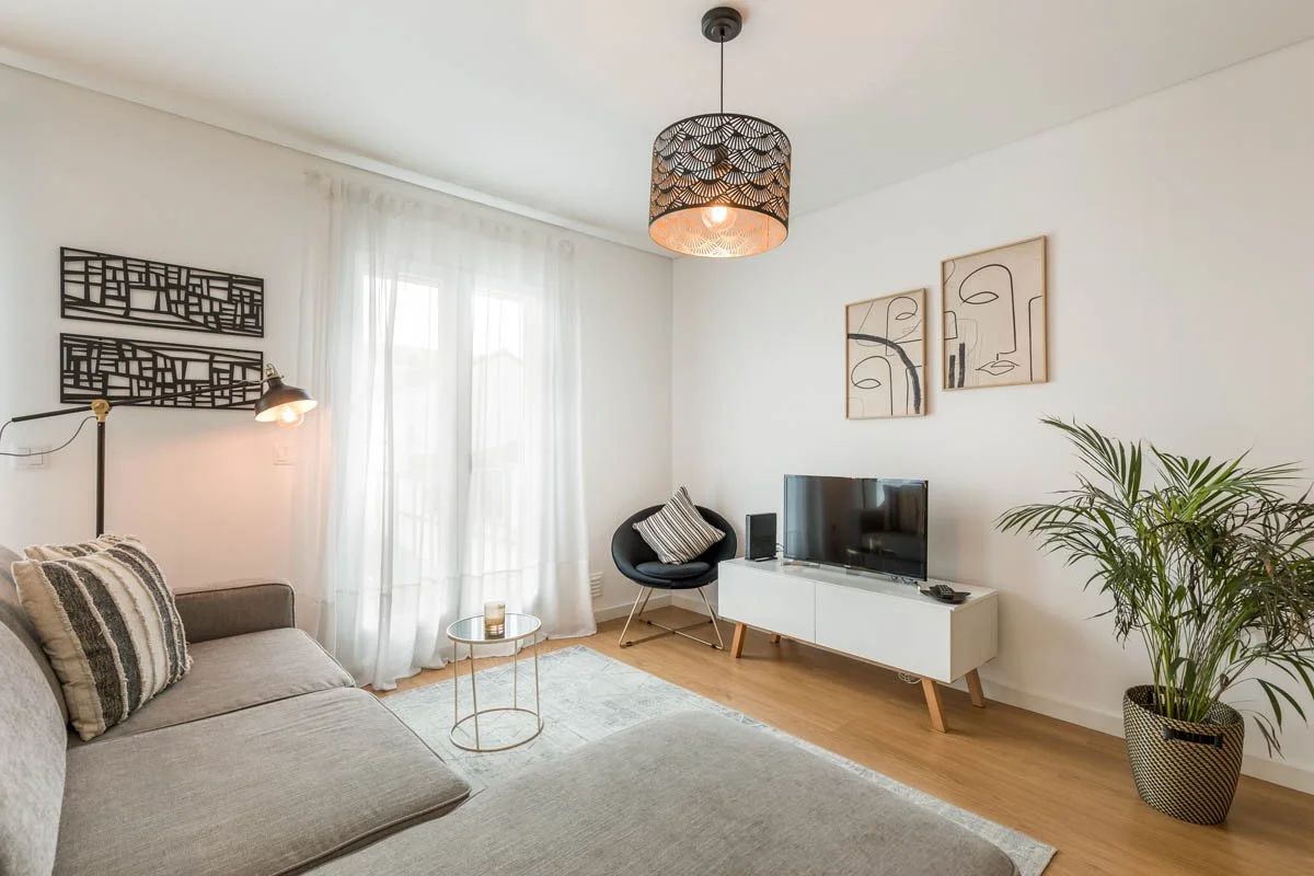 Sala de estar com sofá cinza, parede branca, cortinas brancas, mesa de centro de vidro, televisão em móvel branco, parede decorada com duas obras de arte abstratas, luminária de teto decorativa, luminária de parede ajustável, poltrona preta com almof