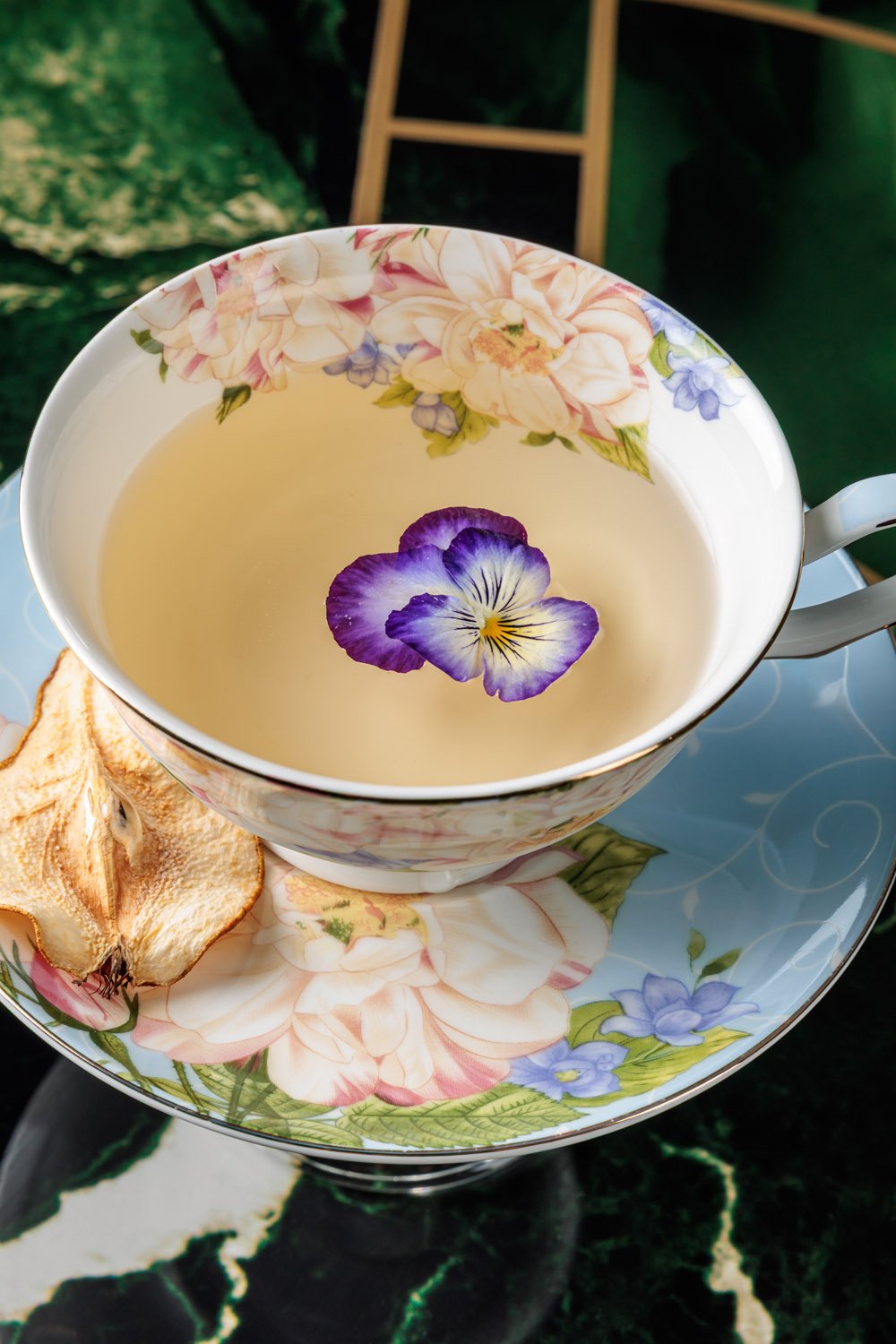 Chá ou infusão em uma xícara decorada com flores, com uma flor violeta na superfície. Acompanhada por uma fatia de maçã assada ou desidratada, sobre um prato também floral.