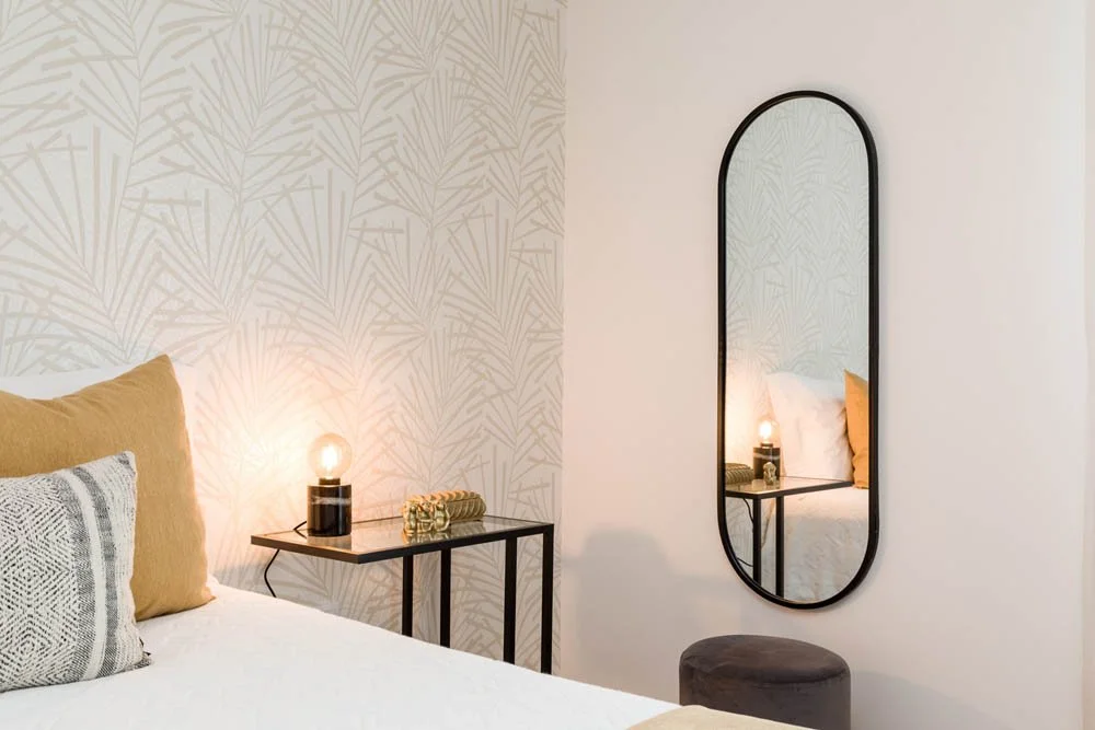 Quarto decorado com papel de parede de padrão tropical, cama com travesseiros amarelos e cinza, espelho de vidro de parede, mesa de cabeceira preta com luminária de vidro e objetos decorativos.