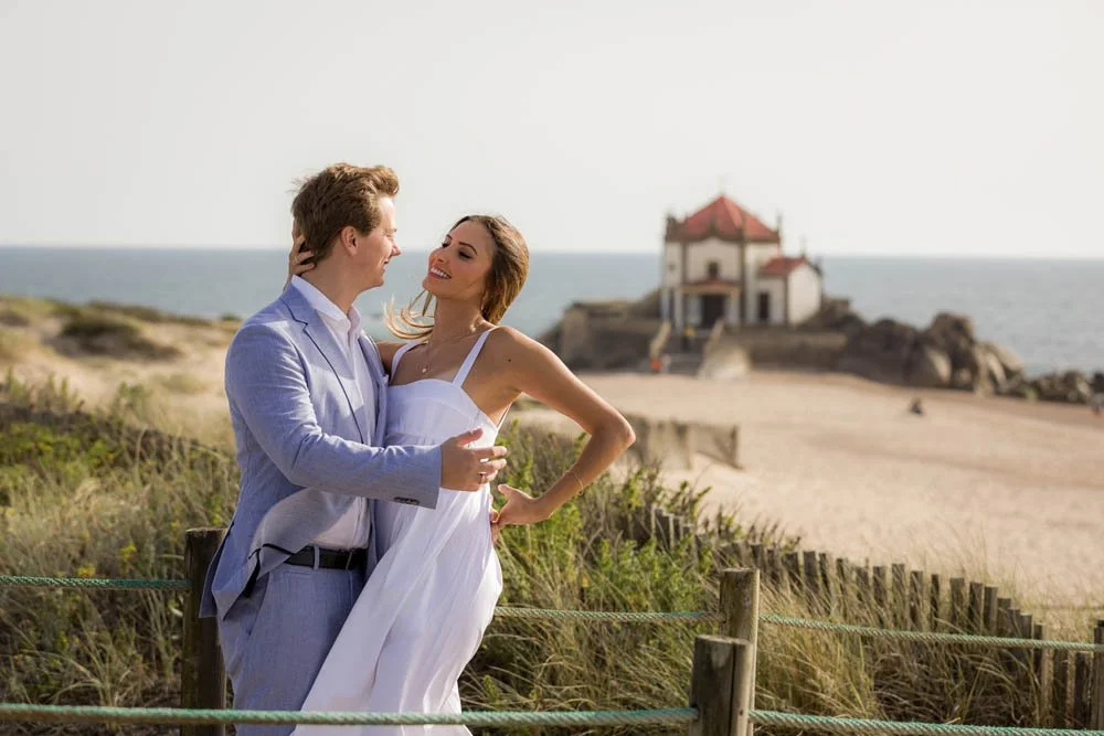 Casal de mãos dadas na praia, com um prédio ao fundo, sob céu claro.