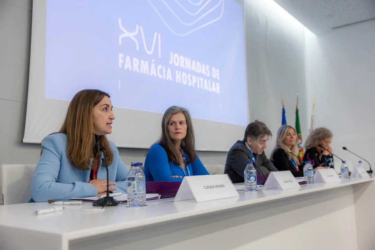 Painel com pessoas sentadas na mesa de uma conferência ou reunião, com fundo com logotipo e texto 'Jornadas de Farmácia Hospitalar'.
