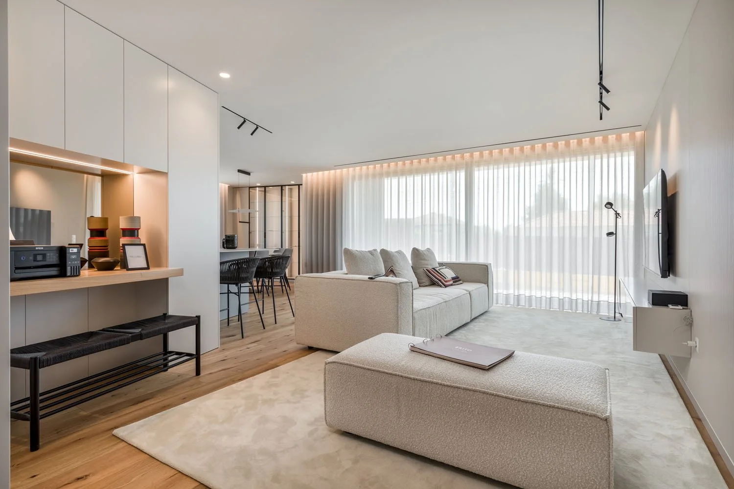 Sala de estar moderna com sofá bege, tapete claro, televisão na parede, cortina de três folhas e iluminação embutida, com espaço para refeições ao fundo.