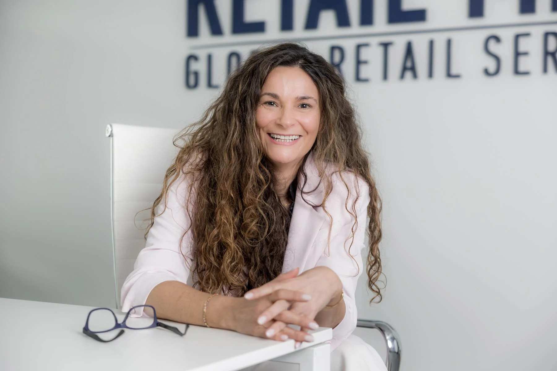 Mulher sorridente com cabelos cacheados longos, usando blazer claro, sentada em uma mesa de escritório, com óculos escuros à sua frente, atrás de uma parede com o texto 'RETAIL' parcialmente visível