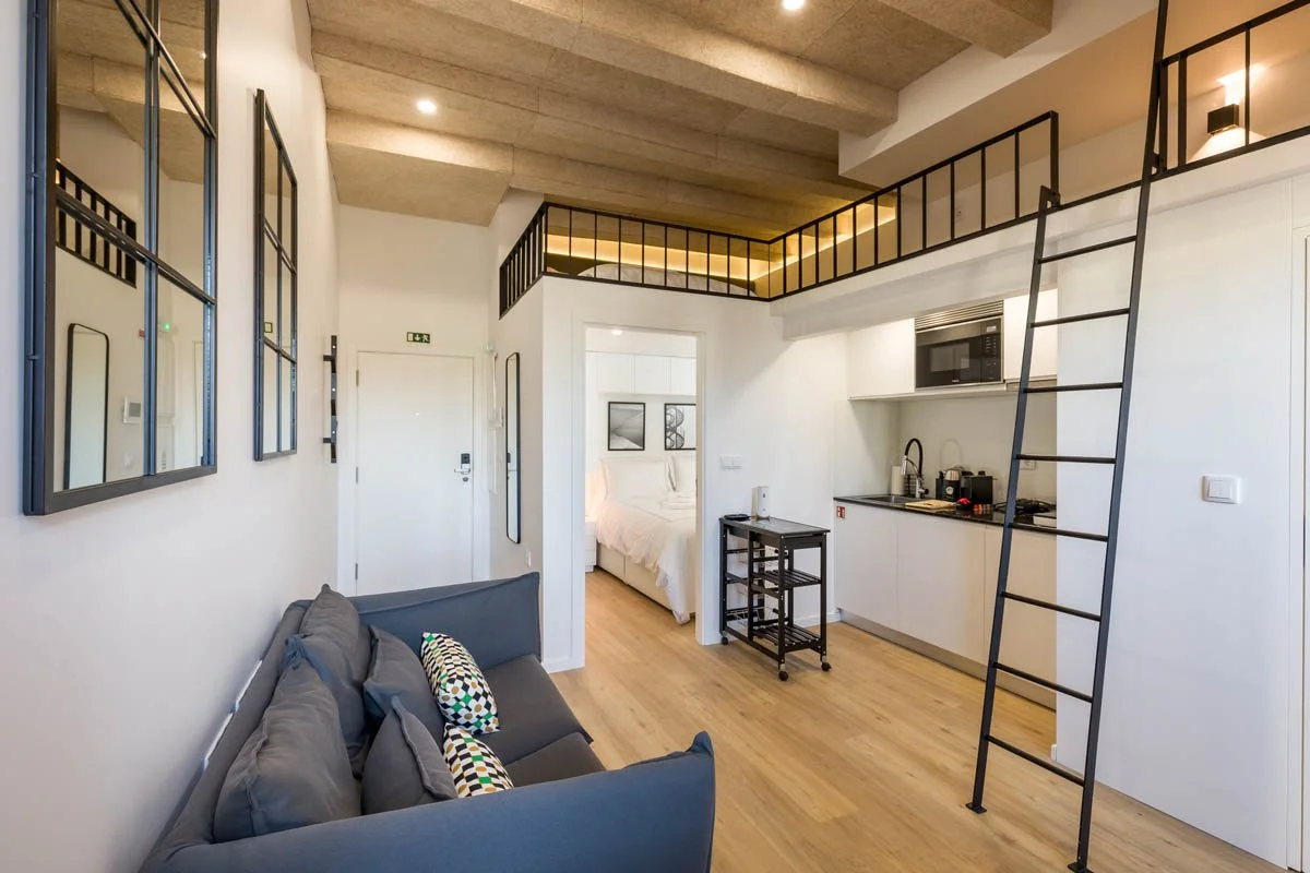 Sala de estar com sofá cinza e almofadas coloridas, vista para um quarto com cama branca, e uma cozinha pequena com micro-ondas, fogão e pia. Uma escada de metal leva ao mezanino com guarda-corpo de ferro. Parede branca e piso de madeira.