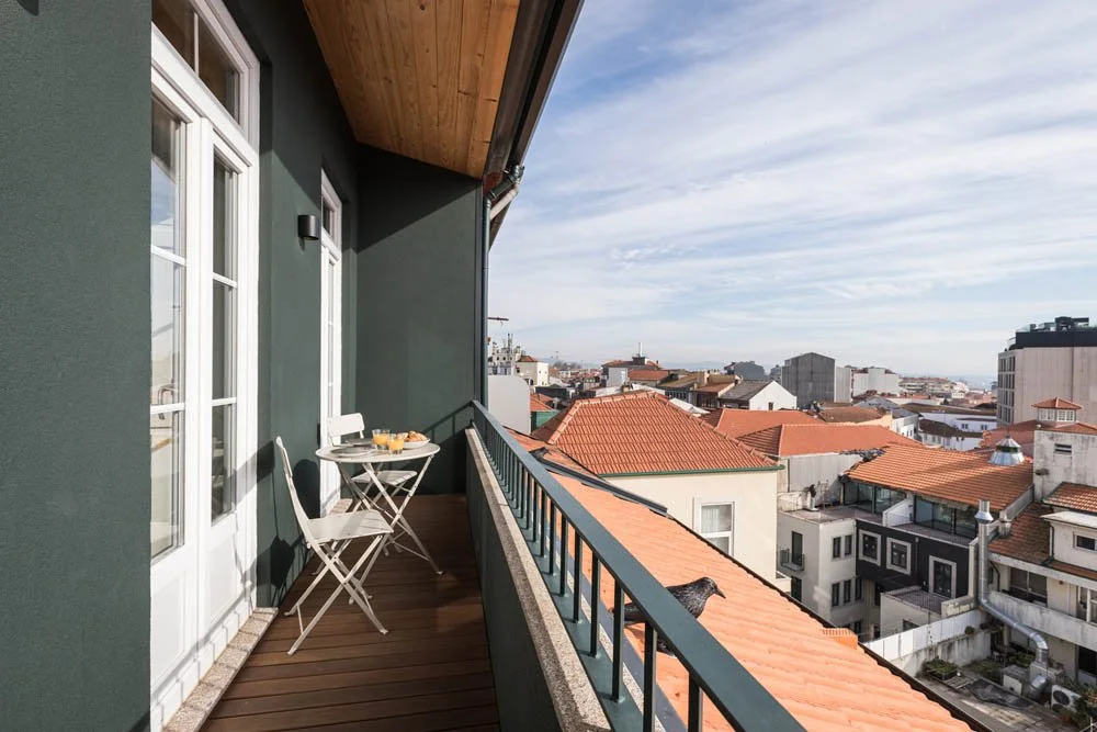 Varanda de apartamento com mesa e duas cadeiras, vista para telhados e prédios da cidade ao fundo.