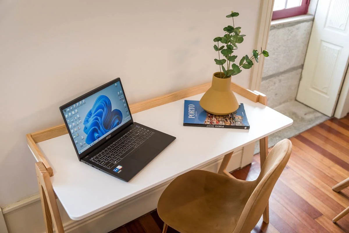 Escritório com mesa branca, cadeira de madeira, laptop ligado, vaso com plantas e livros sobre portugal.