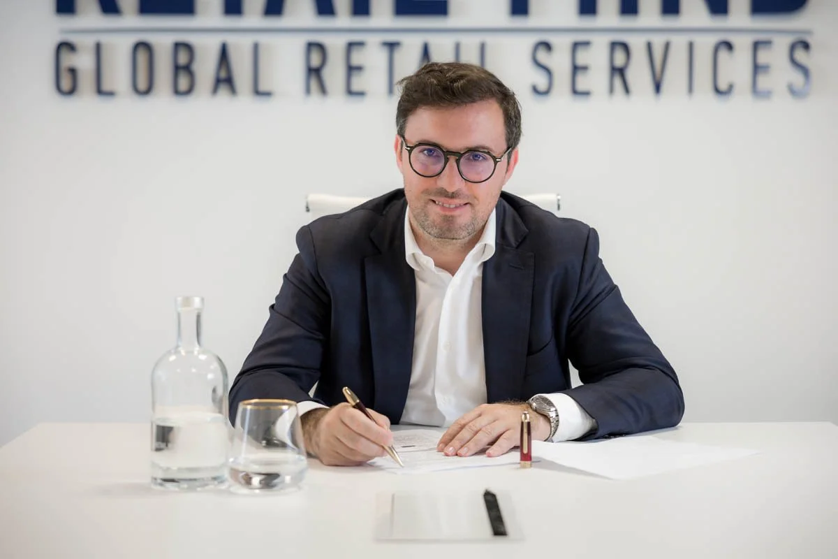 Homem de terno sentado em uma mesa de escritório, segurando uma caneta, com garrafa de água e documentos, atrás um muro com letras de palavras em inglês.