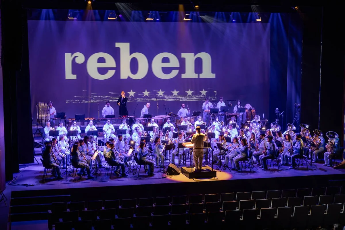 Orquestra de músicos tocando em um palco, com uma pessoa conduzindo, e a palavra 'reben' projetada ao fundo em um telão, com iluminação azulada.