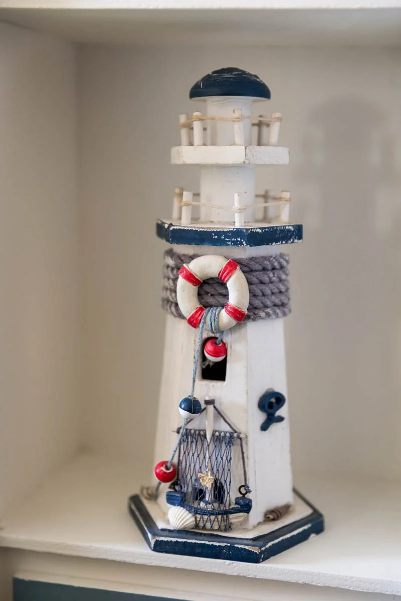 Miniatura de um farol decorativo com detalhes de corda, boias, uma rede, uma âncora, conchas e cores azul e branco, com estilo náutico.
