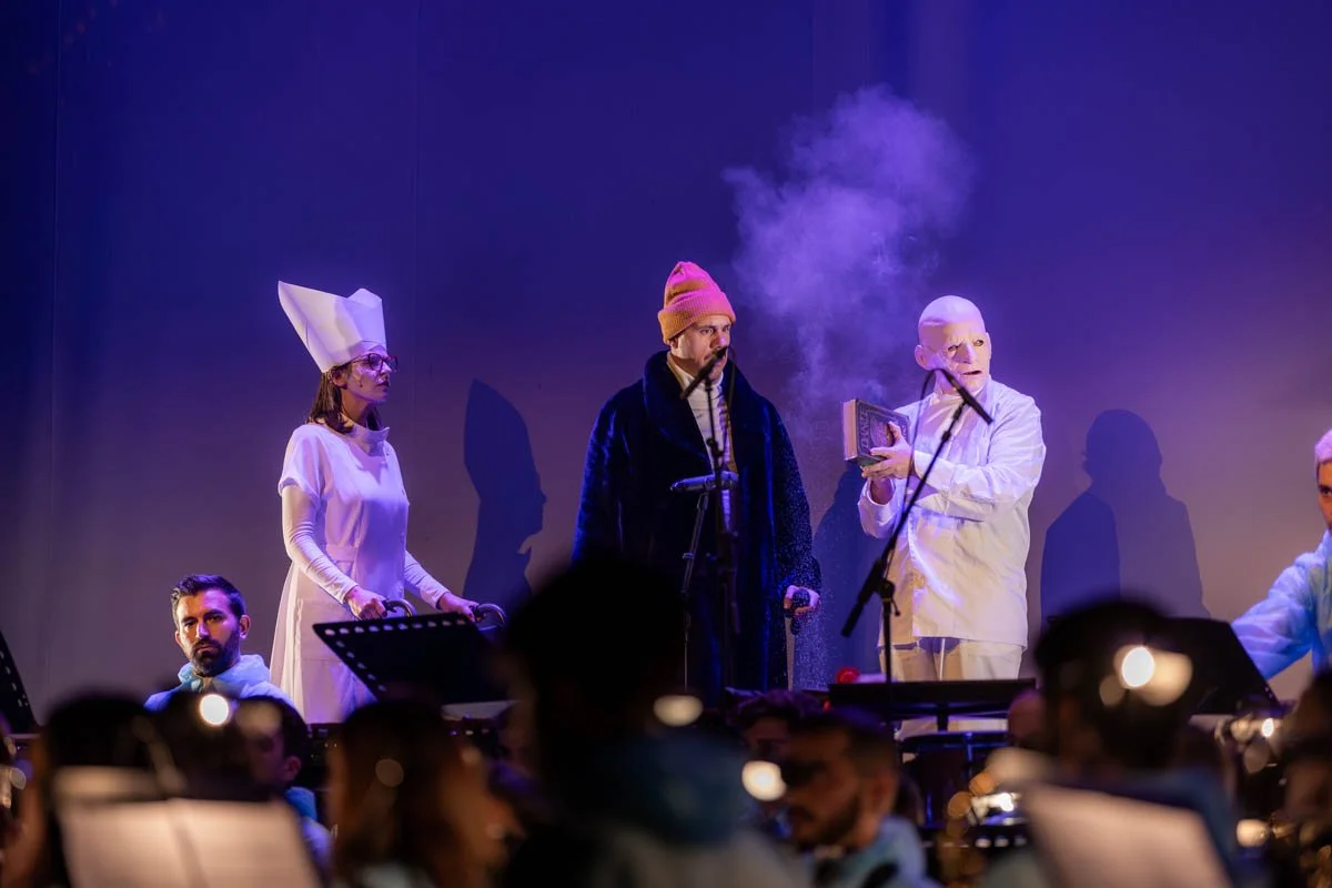 Atores em uma produção teatral com figurinos e máscaras, incluindo uma pessoa com roupa branca, uma com máscara branca, outra com chapéu rosa e uma com chapéu de papelão, em um palco com fumaça e iluminação dramática.