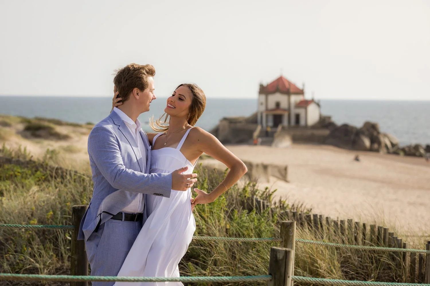 Casal apaixonado na praia, sorrindo e se olhando, com uma igreja ao fundo, durante o pôr do sol.