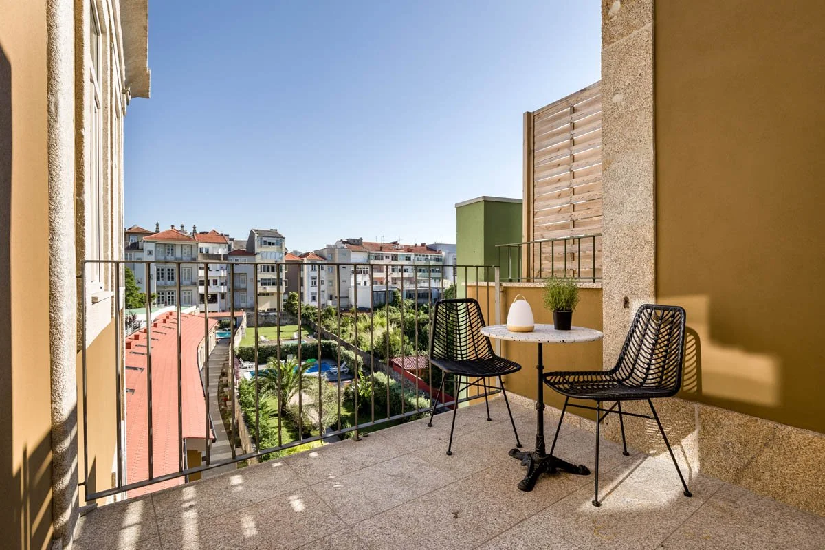 Varanda com duas cadeiras de metal, uma mesa pequena com vaso decorativo e planta, com vista para prédio de apartamentos e jardim ao fundo, em dia ensolarado.