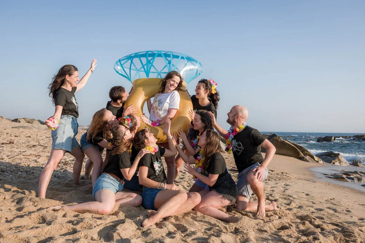 Grupo de amigos celebrando na praia com uma bóia de anel de diamante inflável, alguns usando colares de flores e camisetas pretas, comemorações e risadas sob céu azul.