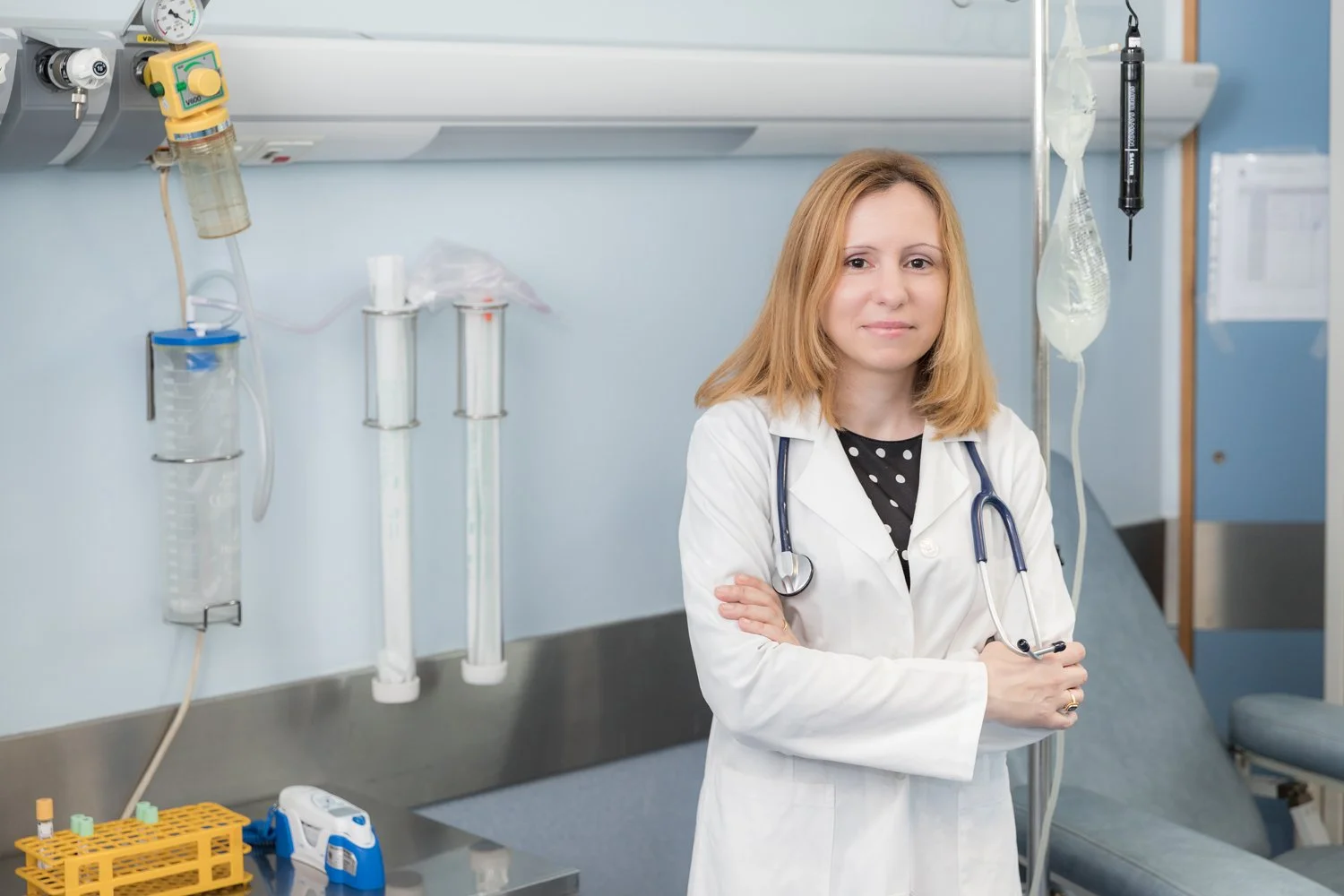 Médica sorridente em ambiente de hospital, vestida com jaleco branco e estetoscópio ao pescoço.