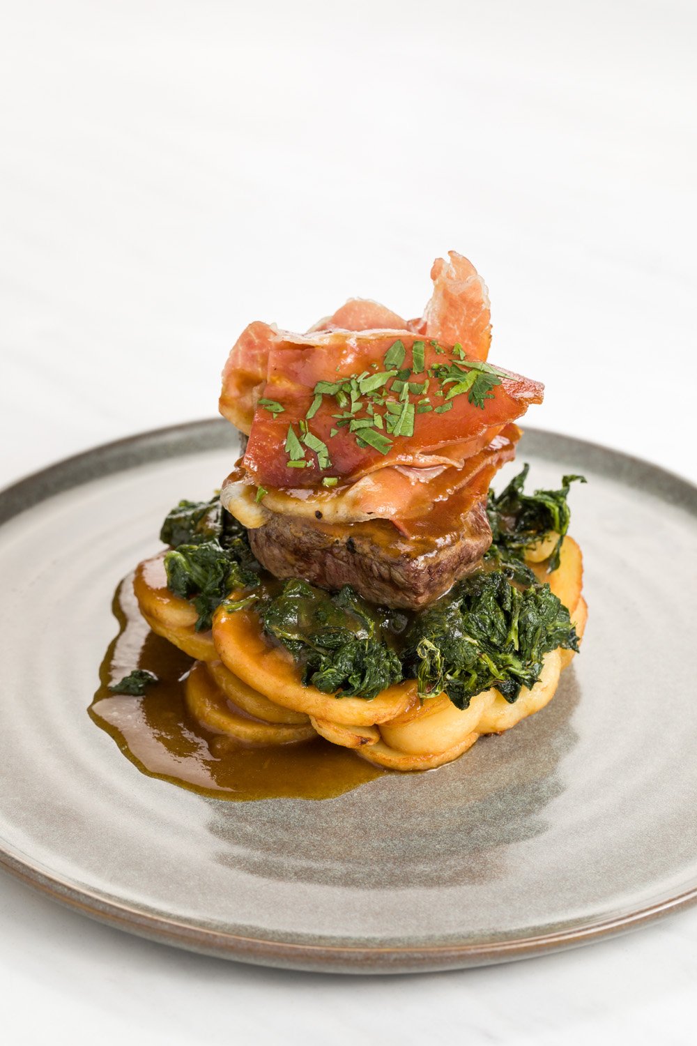 Prato de comida com carne, legumes e folhas verdes, decorado com molho e ervas, servido em um prato cinza