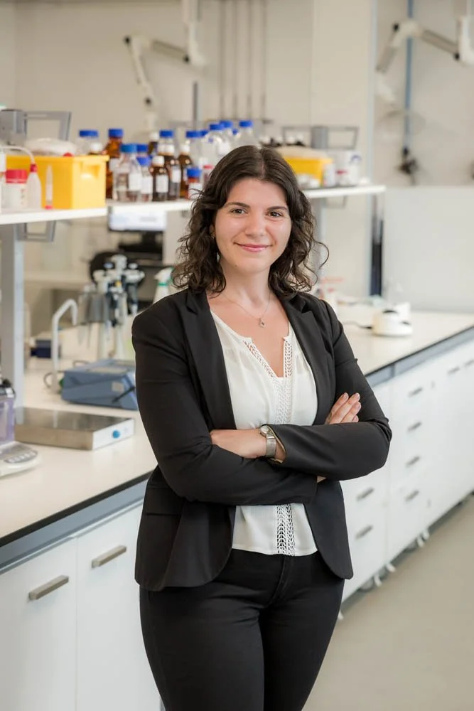Mulher sorridente em roupa formal, de braços cruzados, em laboratório com bancadas, equipamentos e frascos ao fundo.