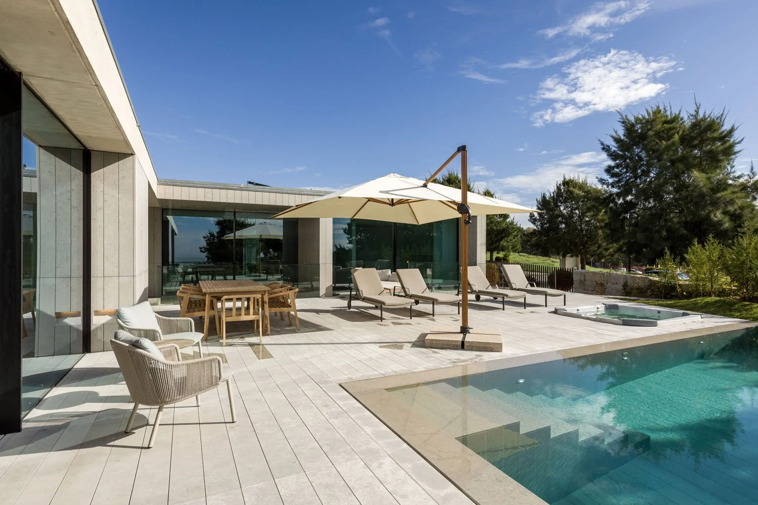 Área externa de uma casa moderna com piscina, espreguiçadeiras, guarda-sol, mesas de jantar e cadeiras, cercada por árvores e céu azul.